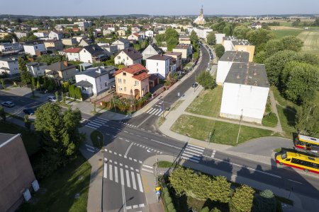 Fragment Ciechanowa widziany z góry. Zdjęcie zrobione przy pomocy drona. Pośrodku, w dolnej połowie fotografii jest skrzyżowanie ulic. Jest tu ciemny asfalt i wyraźnie namalowane linie pasów na przejściach dla pieszych oraz linie wyznaczające pasy ruchu i ścieżki rowerowe. Od skrzyżowania łukiem w górę biegnie ulica Asnyka. Można dostrzec, że ulica ma nową nawierzchnię. Po lewej stronie ulicy Asnyka są domy jednorodzinne, a po prawej budynki wielorodzinne. W oddali widać kościół parafii św. Piotra Apostoła.