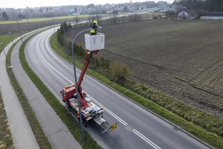 Na zdjęciu fragment drogi w Ciechanowie. Droga biegnie łukiem w prawo w kierunku Osiedla Zachód i wiedzie na widoczne w oddali wzniesienie. Przy jednej z lamp wzdłuż jezdni stoi pojazd z wysięgnikiem. W koszu wysięgnika, uniesionym aż do klosza lampy, stoi pracownik w żółtym kasku i odblaskowej kamizelce.