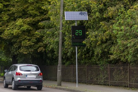 Przy ulicy stoi radarowy wyświetlacz prędkości. Urządzenie mierzy prędkość z jaką nadjeżdża samochód i pokazuje ją na dużym wyświetlaczu. Wyświetlacz przymocowany jest do wysokiego słupka. Urządzenie jest samowystarczalne, jeżeli chodzi o zapotrzebowanie na energię elektryczną. Nad wyświetlaczem znajduje się panel fotowoltaiczny, który przekształca energię słoneczną na elektryczną. Na fotografii Obok wyświetlacza przejechał właśnie samochód osobowy z dopuszczalną prędkością 29 km na godzinę. Liczba wyświetliła się na zielono.