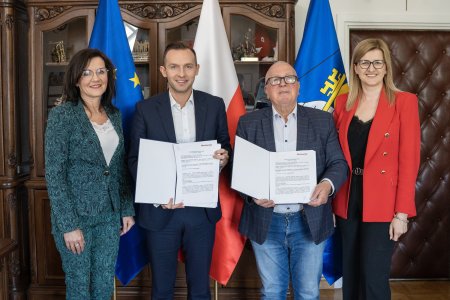 Zdjęcie wykonane w gabinecie prezydenta Krzysztofa Kosińskiego. Na tle flag: unijnej, polskiej biało-czerwonej i flagi z herbem Ciechanowa stoją przedstawiciele Urzędu Miasta i firmy PLAST-BUD, wykonawcy przebudowy ulicy Wiśniowej. Urząd reprezentują: prezydent Krzysztof Kosiński i zastępca prezydenta Iwona Kowalczuk i Luiza Staszewska, kierownik Wydziału Finansowo-Budżetowego. Wykonawcę inwestycji zaś reprezentuje właściciel firmy Kazimierz Olszewski. K. Kosiński i K. Olszewski prezentują egzemplarze podpisanej umowy.