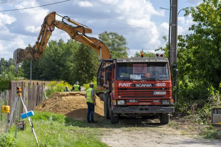 Na środku zdjęcia, w otoczeniu drzew i krzewów na wprost i po prawej oraz prywatnej posesji po lewej, stoi wywrotka. Za wywrotką pracuje koparka, która wysypuje żwir tuż obok powstającej, choć gruntowej jeszcze drogi. W rogu fotografii po lewej stoi teodolit - urządzenie geodezyjne, szeroko stosowane w różnych gałęziach budownictwa, służące do wyznaczania kątów i odchyleń od pionów. Obok wywrotki stoi pracownik w odblaskowej kamizelce. Dwóch pracowników znajduje się bliżej koparki.