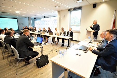 Sala konferencyjna w ratuszu podczas obrad Rady Miasta. Radni i władze miasta siedzą za dwoma długimi stołami, stojącymi równolegle do siebie. Stoły te stoją prostopadle do stołu prezydialnego, który znajduje się na szczycie po prawej. Przy mównicy stoi Zdzisław Dąbrowski, zastępca przewodniczącego Rady Miasta.