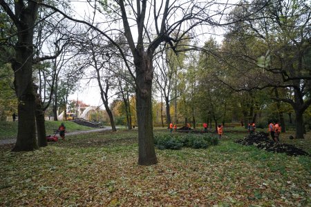 Park J. Dąbrowskiego. Rosną tu wysokie stare drzewa. Niektóre zgubiły już większość liści. W głębi zdjęcia pracownicy zieleni w pomarańczowych kamizelkach sadzą nowe rośliny. Park zyska XIX-wieczny charakter.