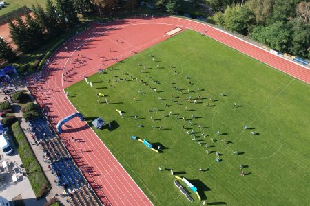 Fragment miejskiego stadionu. Zdjęcie wykonane z góry. Na stadionie znajduje się wielu sportowców. Z wysoka wyglądają jak maleńkie kropeczki. Przy dolnej krawędzi zdjęcia znajduje się podium.