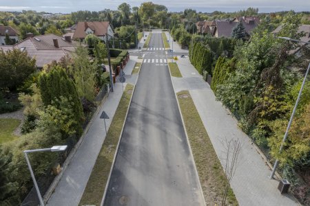 Zdjęcie wykonane z góry. Na środku fotografii, od dołu go góry, biegnie nowa, równa ulica z dwoma przejściami dla pieszych oraz chodnikami i lampami oświetleniowymi po obu stronach. Po bokach zdjęcia, wzdłuż ulicy mieszczą się prywatne posesje. Spośród pięknej zieleni wyglądają spadziste dachy domów.