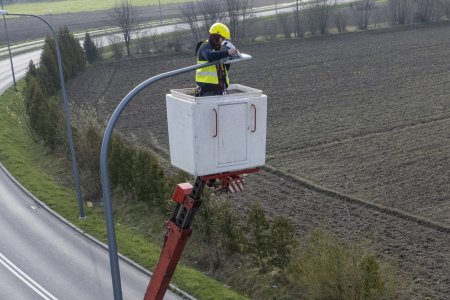 Mężczyzna stojący na wysięgniku wymienia oprawę lampy na energooszczędną. Pracownik ma na sobie odblaskową kamizelkę i jaskrawy ochronny kask. Lampa, przy której pracuje stoi tuż przed zakrętem drogi w prawo pod kątem niemal 90 stopni. Po przeciwległej stronie ulicy znajduje się zaorane pole, a na jego obrzeżu rosną krzewy. 