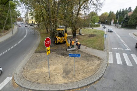 Trwają prace na skwerze. Pracuje tu nieduża żołta koparka, ktora ubija ziemię przymocowanym do ramienia metalowym wałem. W pobliżu pracują ludzie w odblaskowych kamizelkach.