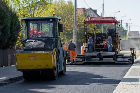 Na niezbyt szerokiej ulicy pracują maszyny do rozściełania i zagęszczania asfaltu. Maszyna do rozściełania asfaltu jest tak szeroka, że układa nawierzchnię od razu na całej szerokości jezdni. Walec jest ponad dwukrotnie węższy. Po obu stronach ulicy są chodniki z brukowej kostki. Po lewej stronie znajduje się rząd prywatnych posesji z jednorodzinnymi domami. Rośnie tu dużo krzewów, których liście zaczynają się wybarwiać na żółto z powodu zbliżającej się zieleni.