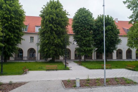 Krzywa Hala sfotografowana od strony skweru na placu Piłsudskiego. Hala ma spadzisty dach w kolorze ceglanym, jasną elewację i charakterystyczne, długie podcienia z wieloma łukami. podcienia.