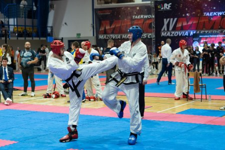 W hali sportowej trwają zawody taekwon-do. Na ścianach hali wiszą wielkie czarno-biało-czerwone banery z napisami: Taekwon-do ITF 15 Mazovia Masters Cup. Na pierwszym planie znajduje się dwóch walczących zawodników w białych dobokach (stroje do ćwiczeń i walki). Na głowach i stopach mają ochraniacze. W tle stoi kilku innych zawodników. Po lewej na krzesłach siedzą sędziowie ubrani w garnitury. W głębi, za barierkami stoją widzowie.