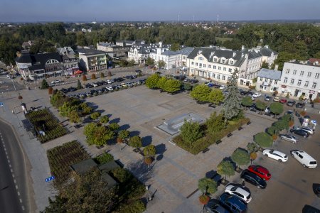 Zdjęcie wykonane z wysoka od południowo wschodniej strony placu przed ratuszem. W kadrze mieści się cały plac Jana Pawła II wraz z zabudowaniami po jego północnej i zachodniej stronie. Zdjęcie objęło także odległe okolice na północny zachód, z porośniętą zielenią Doliną Rzeki Łydyni oraz dużym osiedlem domów jednorodzinnych.