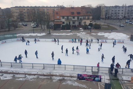 Wielu miłośników łyżew korzystających z miejskiego sezonowego lodowiska przed halą widowiskowo-sportową. Zdjęcie wykonane z budynku hali. W tle ulica 17 Stycznia i budynki po drugiej stronie ulicy.