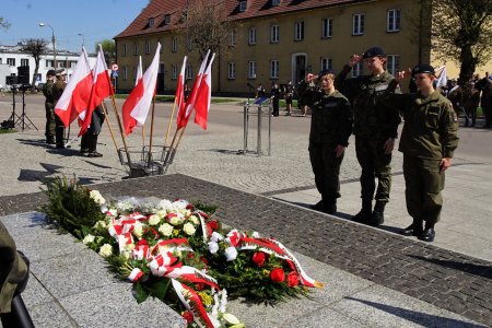 Uroczystości na placu Marszałka Jozefa Piłsudskiego. Trzech żołnierzy (są to kobiety) salutują pod pomnikiem Marszałka. Przed pomnikiem leżą wieńce z białych i czerwonych roż, przewiązane biało-czerwonymi wstęgami. W oddali stoi orkiestra Ciechanowskiej Ochotniczej Straży Pożarnej oraz inni uczestnicy uroczystości.