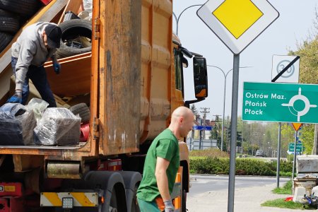 Dwoch mężczyzn zabiera odpady wielkogabarytowe ułożone przed posesjami. Jeden z panow podaje niepotrzebne mieszkańcom rzeczy, a drugi z panow układa je w wielkim kontenerze pomarańczowego pojazdu Przedsiębiorstwa Usług Komunalnych. W kontenerze leżą już stare meble i dużo zużytych opon.