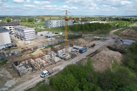 Plac budowy nowego bloku. Zdjęcie wykonane z wysoka, z drona. Z ziemi wystają już prostokątne fundamenty i gdzieniegdzie stoją fragmenty ścian. Obok powstającego budynku stoi wysoki, żołty żuraw. Po lewej stronie znajdują się trzy czterokondygnacyjne bloki. Jeden z nich ma rusztowania dookoła elewacji. W oddali na wprost też stoi niewysoki budynek wielorodzinny. Po prawej stronie powstającego bloku leży hałda żwiru, a dalej w prawo znajduje się teren porośnięty krzewami i młodymi drzewami.