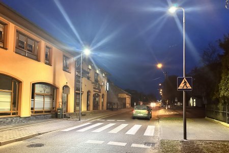 Wyraźnie oświetlone przejście dla pieszych na ul. Rzeczkowskiej. Lampy stoją po jednej i drugiej stronie ulicy. Jedna z prawej, druga z lewej strony przejścia dla pieszych. Po lewej stronie ulicy znajduje się długi żółty budynek Zakładu Ubezpieczeń Społecznych.