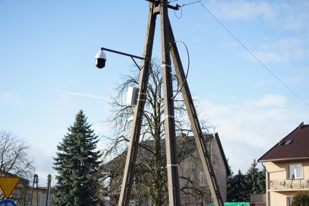 Kamera monitoringu zamontowana na słupie linii elektrycznej. W tle zdjęcia znajdują się dachy domów jednorodzinnych.