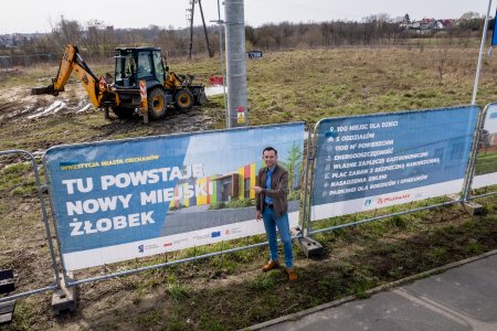 Teren, na ktorym powstanie nowy żłobek. Miejsce to jest otoczone ogrodzeniem z metalowych modułow. Na trzech elementach ogrodzenia wiszą banery z informacjami o inwestycji. Przy pierwszym banerze z ilustracją przyszłego przedszkola stoi prezydent Ciechanowa Krzysztof Kosiński. Prezydent wskazuje grafikę dłonią.