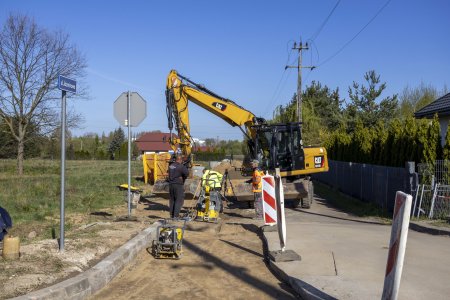 Trwa budowa lewej połowy jezdni ulicy Jaśminowej. Drugą połową można przejechać autem. Wzdłuż remontowanej części drogi układane są krawężniki. Pracuje tam dwoch mężczyzn, ktorzy mają do pomocy żołtą koparkę. Mężczyźni ubijają podłoże korzystając z zagęszczarek gruntu. Operator podaje ciężkie krawężniki na łysze koparki. Po lewej stronie drogi jest teren porośnięty trawą, w oddali stoją domy. Po prawej stronie znajduje się ogrodzona posesja. Za ogrodzeniem rośnie żywopłot z krzewow.