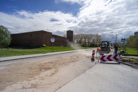 Roboty na ulicy Zamkowej. Zdjęcie wykonane od strony ulicy Parkowej. Na prawym pasie ruchu pracuje trzech mężczyzn w odblaskowych kamizelkach. Dalej stoi ciągnik z łychą. Na zdjęciu, po lewej stronie ulicy Zamkowej stoi Zamek Książąt Mazowieckich.