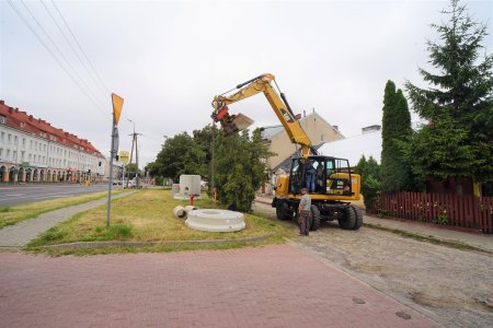 Po lewej stronie zdjęcia znajduje się Hala Pułtuska. Przed halą biegnie głowna ul. Pułtuska. Dolny odcinek ul. Pułtuskiej mieści się po prawej stronie fotografii. Pracuje tu duża żołta koparka. Obok maszyny stoi mężczyzna. Na trawniku wzdłuż drogi ułożono materiały budowlane. 