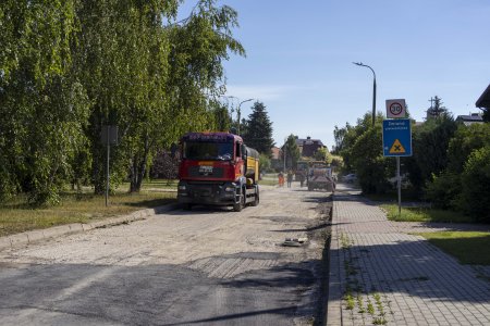 Trwa budowa ul. Baczyńskiego. Droga biegnie wśrod zieleni. Jezdnia jest pozbawiona nawierzchni. Na pierwszym planie zdjęcia znajduje się duży pojazd ciężarowy. W oddali w obłoku pyłu pracuje kilku mężczyzn. Stoją tam też dwa pojazdy firmy budowlanej.