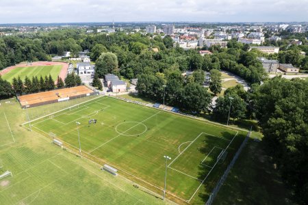 Zmodernizowane boisko piłkarskie, mieszczące się w kompleksie sportowym obok miejskiego stadionu. Cały kompleks sportowy sfotografowany z wysoka. Wokoło boiska i stadionu rośnie dużo starych, pięknych drzew. W głębi fotografii panorama miasta. Na boisku trwa trening.