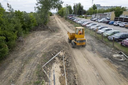 Po wytyczonym szlaku, jakim pobiegnie nowa droga jedzie nieduży żołty ciągnik z przyczepką. Droga jest długa i piaszczysta. Po lewej rosną drzewa i krzewy, a po prawej znajduje się ogrodzony parking. Stoi tu wiele osobowych aut. Na dole kadru, po lewej stronie drogi białą taśmą otoczono miejsce, w ktorym odkopano dwa podłużne elementy. Wyglądają jak metalowe rury.