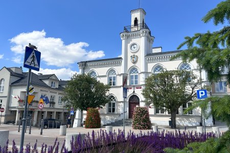 Ciechanowski ratusz przy pięknej, letniej pogodzie. Zabytkowy budynek ma jasne ściany, ozdobne gzymsy i wieżyczkę z zegarem oraz balustradą. Na szczycie znajduje się iglica z flagą Ciechanowa. Przed ratuszem rosną dwa drzewiaste głogi, stoją tu wysokie kwietniki z surfiniami. Obok, po lewej znajduje się fragment drugiego budynku zajmowanego przez Urząd Miasta.