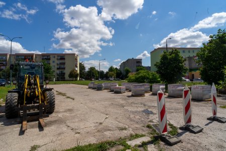 Duży plac budowy nowego parkingu. Po lewej stoi ciągnik z podnośnikiem widłowym. Pojazd ten dostarczył na plac 14 palet z brukową kostką. Palety stoją po prawej stronie zdjęcia. Wzdłuż prawej krawędzi placu rosną spore drzewa. Wjazd na teren budowy zamykają biało czerwone słupki. W tle fotografii stoi kilka pięciokondygnacyjnych blokow.