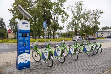 Stacja Ciechanowskiego Roweru Miejskiego IV generacji, zlokalizowana przy dworcu kolejowym. Rowery ustawione rządem w stojakach mają kolor jasnozielony. Oznaczenia na tabliczkach przypiętych po obu stronach bagażnikow są w kolorach białym i niebieskim. W tle stacji rowerowej znajduje się skwer z fontanną, a w głębi po prawej stoi budynek dworca.