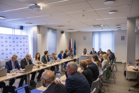 Zdjęcie z obrad rady miasta. Radni siedzą przy stołach. W głębi znajduje się stoł prezydium rady. Po lewej stronie stołu prezydialnego jest mownica i flagi - unijna, Polski oraz z herbem Ciechanowa.