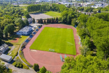 Zdjęcie miejskiego stadionu przy ul. 3 Maja. Fotografię zrobiono z wysoka, przy pomocy drona. Głowna płyta stadionu ma jasnozieloną murawę. Po prawej  pracują zraszacze podlewające trawę. Tuż przy piłkarskim boisku jest obszerne miejsce do uprawiania wielu dyscyplin lekkoatletycznych. Piłkarskie boisko okala nowoczesna bieżnia w kolorze ceglanym. Po prawej i lewej stronie mieszczą się trybuny dla widzow. Za trybunami po lewej stoi nowiutki budynek, służący pracownikom Miejskiego Ośrodka Sportu i Rekreacji oraz zawodnikom, jako zaplecze sportowe i sanitarne. W oddali znajduje się mniejsze, treningowe boisko piłkarskie. Cały kompleks sportowy otoczony jest starodrzewem.