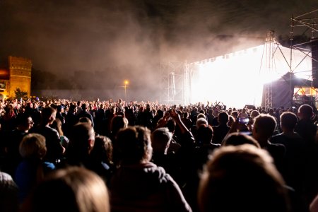 Zdjęcie wykonane na zamkowych błoniach po zmierzchu. Po prawej, na scenie trwa koncert. Bardzo jasne sceniczne oświetlenie i unoszący się dym powodują, że na fotografii nie widać występującego artysty. Przed sceną bawi się bardzo wielu ludzi. Widzowie mają uniesione ręce. Po lewej stronie zdjęcia znajduje się oświetlony fragment baszty Zamku Książąt Mazowieckich.