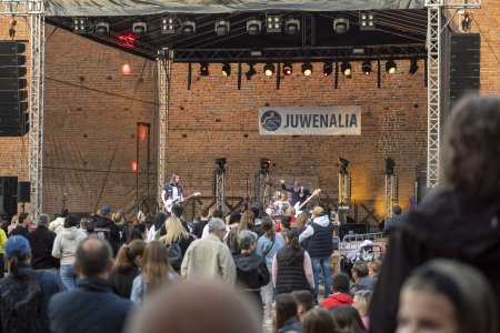 Plenerowa scena z rządami scenicznych świateł na rampie pod zadaszeniem. Scena jest ustawiona pod jedną z ceglanych ścian na dziedzińcu Zamku Książąt Mazowieckich w Ciechanowie. Na scenie gra zespoł.  Na froncie grają dwaj gitarzyści. Za ich plecami rytm wybija perkusista. W głębi sceny znajdują się sceniczne lampy na zamontowane na wspornikach. Na ścianie za sceną wisi baner z logo Państwowej Akademii Nauk Stosowanych i dużym napisem Juwenalia. Przed sceną stoi wielu ludzi.