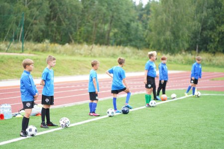 Trening piłkarski. Wzdłuż bocznej białej linii boiska stoi kilku chłopcow. Ubrani są w piłkarskie stroje: niebieskie koszulki i ciemne spodenki lub długie spodnie. Każdy z chłopcow przy nodze ma piłkę. Obok piłkarskiego boiska znajduje się bieżnia.