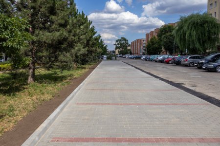 Długi pas parkingowy. Nawierzchnię zbudowano z szarej brukowej kostki. Linie dzielące miejsca parkingowe powstały z dwoch rzędow kostki w kolorze cegły. Po przeciwnej stronie drogi, przy ktorej jest nowy parking, stoi bardzo długi rząd osobowych aut. Po obu stronach drogi i parkingow rosną drzewa. Po prawej, zza drzew wyłania się kilka wielorodzinnych blokow.