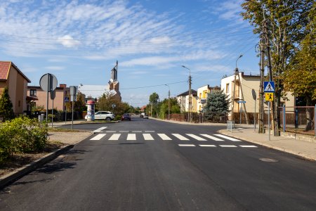 Nowy asfalt na ul. Wyspiańskiego.. Ulica jest szeroka. Nawierzchnia ulicy czarna. Namalowano tu pasy przejścia dla pieszych. Po obu stronach ulicy są zabudowania, a na wprost kościoł z wysoką wieżą.