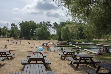 Plaża na Krubinie. Po lewej stronie zdęcia piasek i plażowicze, po prawej zbiornik wodny z prostokątnym pomostem. Dookoła dużo drzew i krzewow.