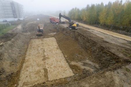 Plac budowy nowego budynku wielorodzinnego. Pracują tu dwie koparki i dwie wywrotki. Powstaje wielki prostokątny wykop pod budowę bloku. Jedna z koparek pracuje na dnie wykopu, druga, stojąc na krawędzi, poszerza jego brzegi. Jest mgła. Po lewej stronie zdjęcia we mgle stoi blok. Po prawej rośnie brzozowy lasek w jesiennych barwach.