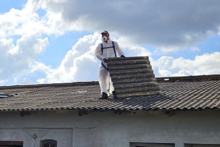 Mężczyzna w białym uniformie i z maską na twarzy demontuje płyty faliste z dachu budynku.