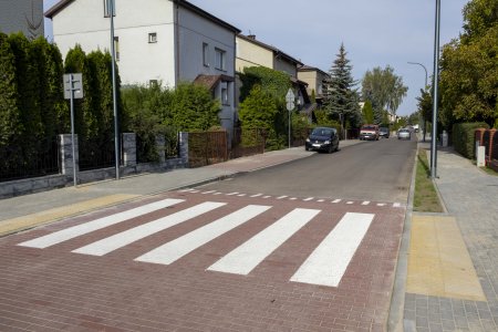 Rowna, wyremontowana ulica. Na pierwszym planie zdjęcia znajduje się tak zwane wyniesione przejście dla pieszych. Zostało zbudowane z czerwonej brukowej kostki i, podobnie jak progi zwalniające, znajduje się nieco powyżej poziomu jezdni. Kierowcy muszą zwalniać przed przejściem dla pieszych. Po obu stronach ulicy znajdują się chodniki, lampy uliczne i prywatne posesje. Rośnie tu dużo drzew i krzewow. W głębi fotografii stoi kilka osobowych aut.