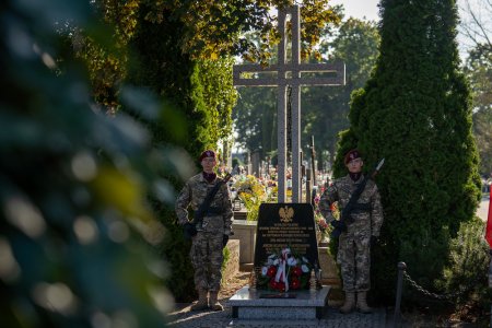 Warta honorowa przy krzyżu upamiętniającym poległych i pomordowanych na Wschodzie. Pod krzyżem leży wiązanka biało-czerwonych kwiatow. Krzyż z pamiątkową tablicą oraz pełniących wartę żołnierzy sfotografowano zza gałęzi drzewa. Nadaje to zdjęciu nieco tajemniczy charakter.