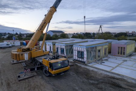 Plac budowy nowego przedszkola. Po lewej stoi wielki żołty dźwig. Po prawej, na utwardzonym podłożu stoją moduły przedszkolnego budynku. Wyglądają jak prostokątne kontenery. Są jeszcze zapakowane.