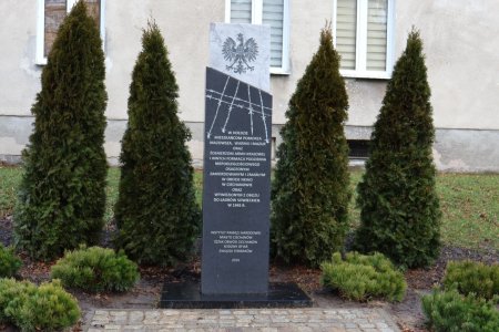 Wysoki prostokątny monument. W gornej, jasnoszarej części umieszczono herb Rzeczypospolitej Polskiej. Poniżej, na ciemnej, grafitowej powierzchni znajduje się biała inskrypcja. Nad napisem wyryto rysunek obozowych kolczastych drutow.